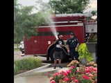 A citizen is standing in front of a fire truck with a fire fighter while spraying the fire hose.