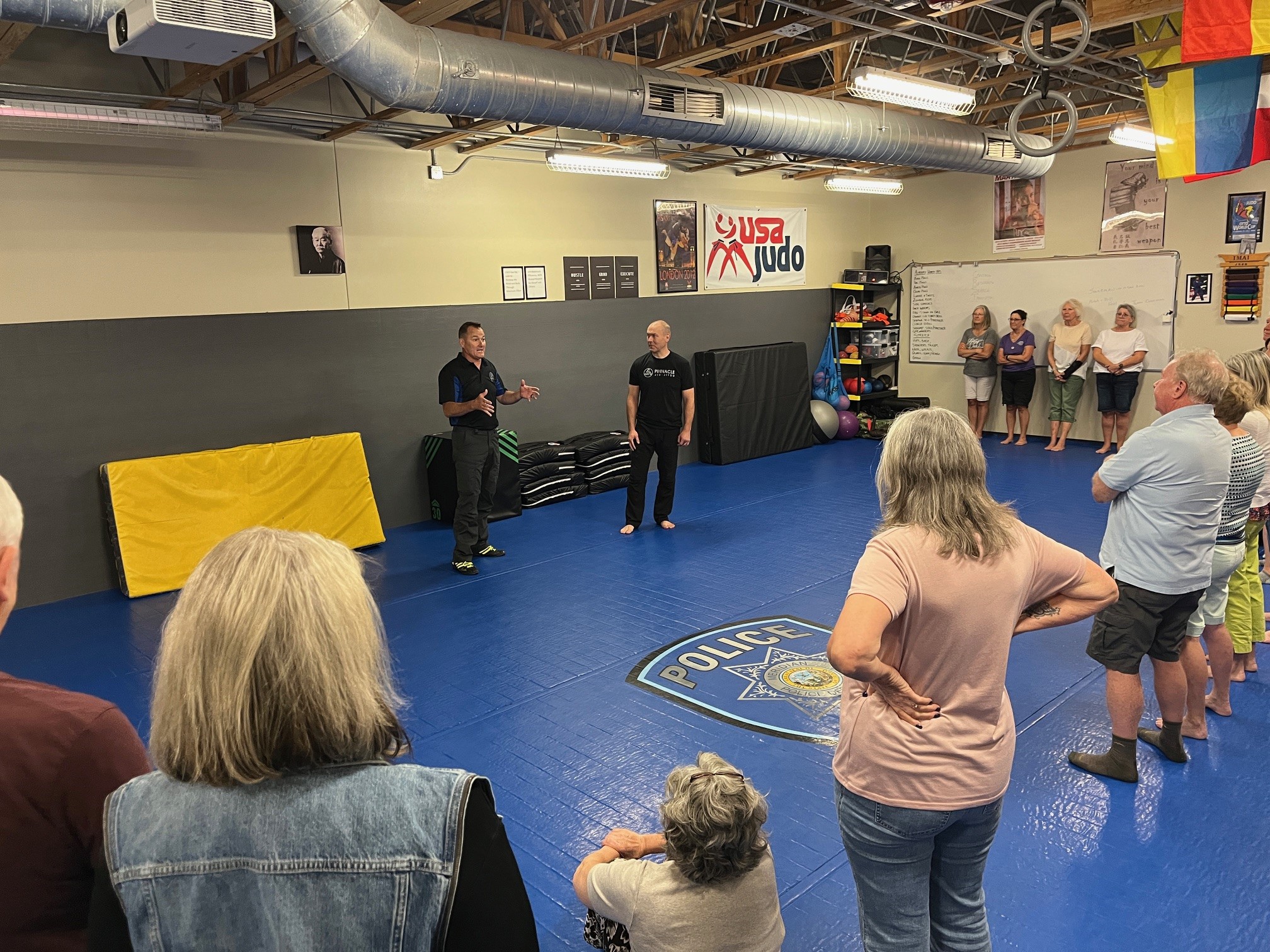 People standing around on blue mat listening to instructors.